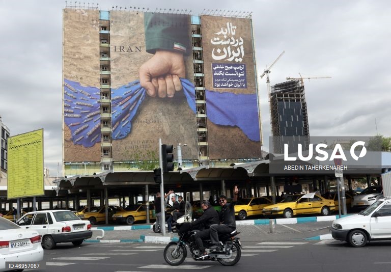 Iranianos passam por um cartaz publicitário de grandes dimensões referente ao Estreito de Ormuz, com a inscrição em persa «Para sempre nas mãos do Irão», numa praça em Teerão, Irão. As tensões entre os Estados Unidos e o Irão continuam, enquanto o Estreito de Ormuz permanece bloqueado.