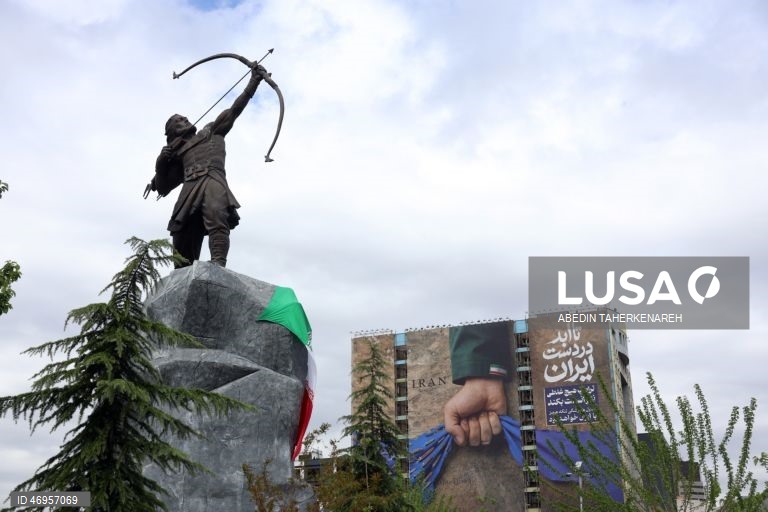 Iranianos passam por um cartaz publicitário de grandes dimensões referente ao Estreito de Ormuz, com a inscrição em persa «Para sempre nas mãos do Irão», numa praça em Teerão, Irão. As tensões entre os Estados Unidos e o Irão continuam, enquanto o Estreito de Ormuz permanece bloqueado.