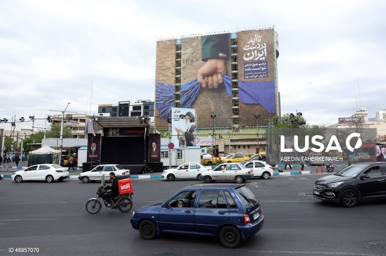 Iranianos passam por um cartaz publicitário de grandes dimensões referente ao Estreito de Ormuz, com a inscrição em persa «Para sempre nas mãos do Irão», numa praça em Teerão, Irão. As tensões entre os Estados Unidos e o Irão continuam, enquanto o Estreito de Ormuz permanece bloqueado.