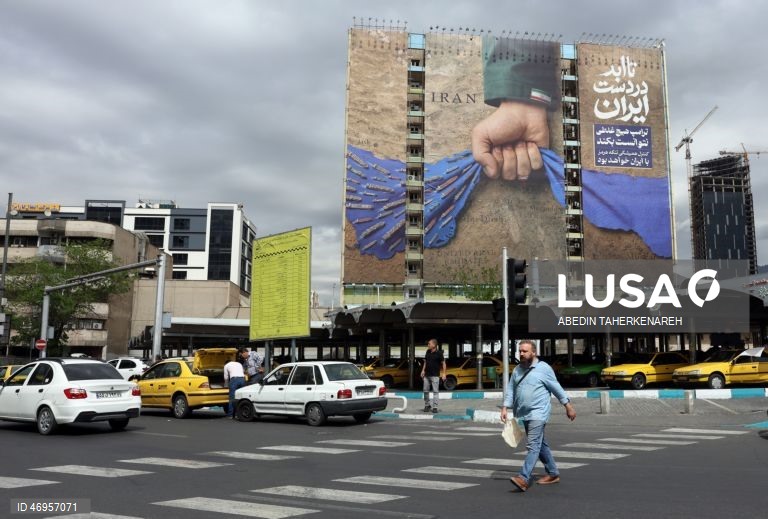 Iranianos passam por um cartaz publicitário de grandes dimensões referente ao Estreito de Ormuz, com a inscrição em persa «Para sempre nas mãos do Irão», numa praça em Teerão, Irão. As tensões entre os Estados Unidos e o Irão continuam, enquanto o Estreito de Ormuz permanece bloqueado.