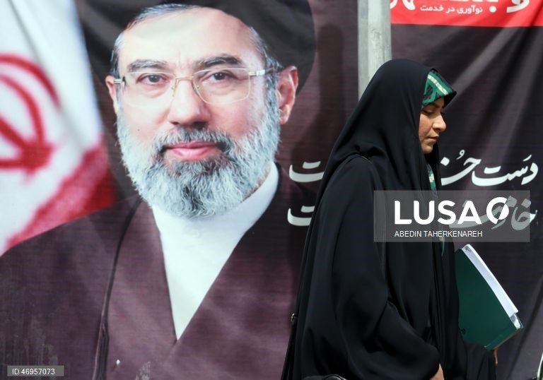 Iranianos passam por um cartaz publicitário de grandes dimensões referente ao Estreito de Ormuz, com a inscrição em persa «Para sempre nas mãos do Irão», numa praça em Teerão, Irão. As tensões entre os Estados Unidos e o Irão continuam, enquanto o Estreito de Ormuz permanece bloqueado.