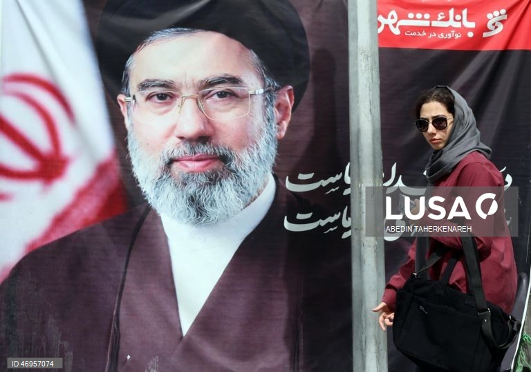 Iranianos passam por um cartaz publicitário de grandes dimensões referente ao Estreito de Ormuz, com a inscrição em persa «Para sempre nas mãos do Irão», numa praça em Teerão, Irão. As tensões entre os Estados Unidos e o Irão continuam, enquanto o Estreito de Ormuz permanece bloqueado.