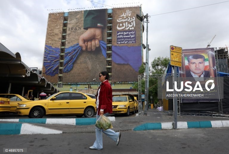 Iranianos passam por um cartaz publicitário de grandes dimensões referente ao Estreito de Ormuz, com a inscrição em persa «Para sempre nas mãos do Irão», numa praça em Teerão, Irão. As tensões entre os Estados Unidos e o Irão continuam, enquanto o Estreito de Ormuz permanece bloqueado.