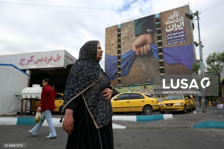 Iranianos passam por um cartaz publicitário de grandes dimensões referente ao Estreito de Ormuz, com a inscrição em persa «Para sempre nas mãos do Irão», numa praça em Teerão, Irão. As tensões entre os Estados Unidos e o Irão continuam, enquanto o Estreito de Ormuz permanece bloqueado.
