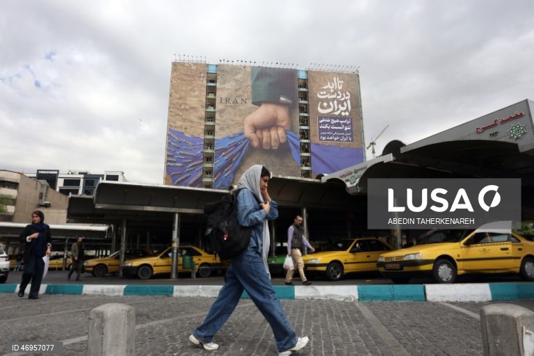 Iranianos passam por um cartaz publicitário de grandes dimensões referente ao Estreito de Ormuz, com a inscrição em persa «Para sempre nas mãos do Irão», numa praça em Teerão, Irão. As tensões entre os Estados Unidos e o Irão continuam, enquanto o Estreito de Ormuz permanece bloqueado.